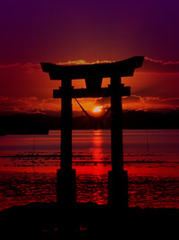A Japanese torii stands before a lake, the sun setting in the background.